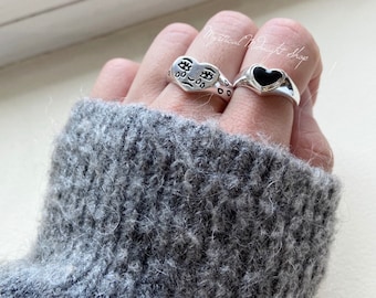 May include: Two silver heart-shaped rings. One ring has a black enamel heart and the other has a face with a sad expression.