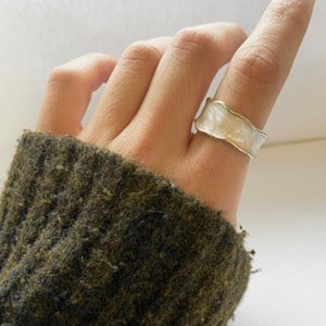 Può includere: Un anello bianco con una fascia dorata e un design ondulato e testurizzato. L'anello è su un dito.