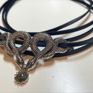 May include: A black leather cord necklace featuring a silver-toned metal pendant shaped like two intertwined snakes. The snakes are textured with a scale-like pattern and meet at the center, where a small, round, dark grey stone is suspended.