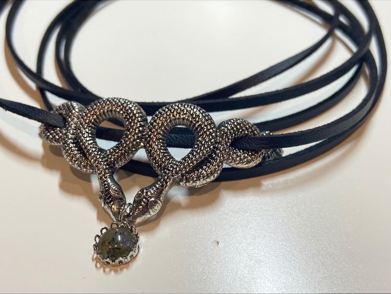 May include: A black leather cord necklace featuring a silver-toned metal pendant shaped like two intertwined snakes. The snakes are textured with a scale-like pattern and meet at the center, where a small, round, dark grey stone is suspended.