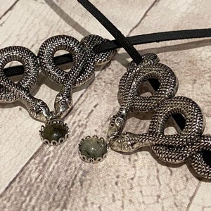 May include: Two silver-toned snake pendants threaded onto a black cord. Each snake is coiled in a circular shape with textured scales and holds a small, round, dark green stone in a scalloped metal setting between its head and tail.