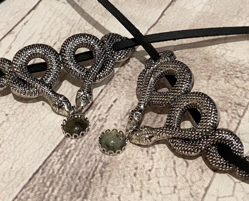 May include: Two silver-toned snake pendants threaded onto a black cord. Each snake is coiled in a circular shape with textured scales and holds a small, round, dark green stone in a scalloped metal setting between its head and tail.