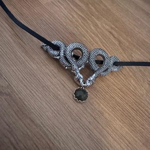 May include: A necklace featuring a black cord and a silver-toned metal pendant shaped like two intertwined snakes. The snakes' heads meet at the bottom, holding a small, round, dark green gemstone in a scalloped silver setting between them.