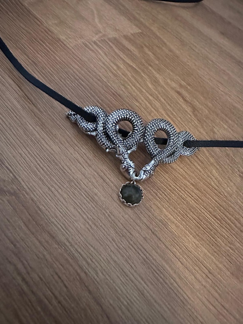 May include: A necklace featuring a black cord and a silver-toned metal pendant shaped like two intertwined snakes. The snakes' heads meet at the bottom, holding a small, round, dark green gemstone in a scalloped silver setting between them.