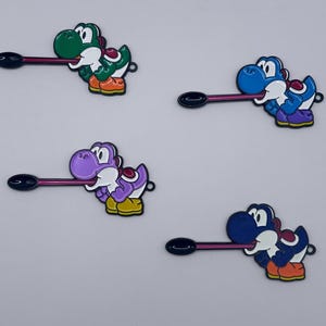 May include: Four colourful Yoshi character pins. Each pin features a different colour Yoshi with a black spoon-like object extending from its mouth. The Yoshis are green, blue, purple, and dark blue, with white, orange, and yellow accents.