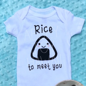 May include: White baby onesie with a black and white illustration of a rice ball with a smiling face. The text "Rice to meet you" is printed below the rice ball. A real rice ball is shown in the lower right corner of the image.