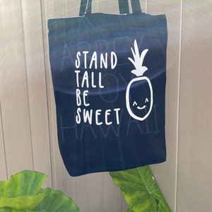 May include: A dark blue tote bag with white text that reads "Stand Tall Be Sweet" and a white pineapple graphic with a smiling face.