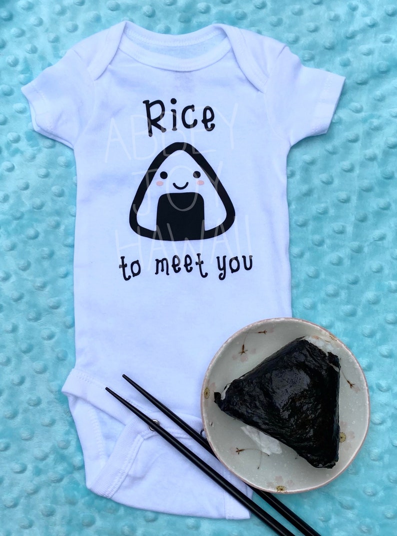 May include: White baby onesie with a black and white illustration of a rice ball with a smiling face. The text "Rice to meet you" is printed below the rice ball. A real rice ball is shown in the lower right corner of the image.