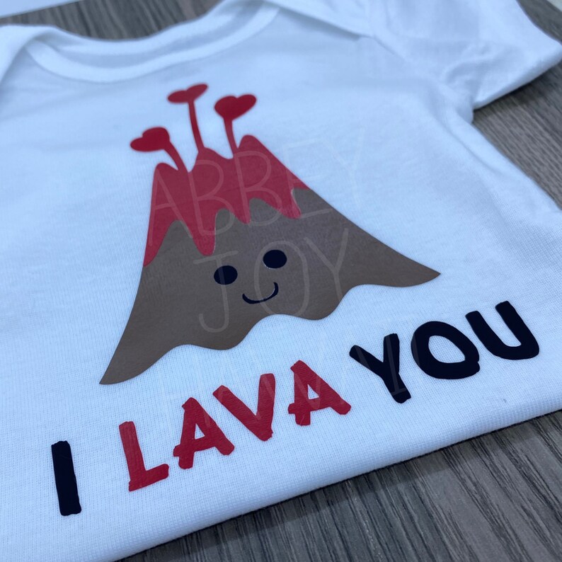 May include: White bodysuit with a brown and red volcano graphic with red hearts on top. The text "I lava you" is written in black below the volcano.