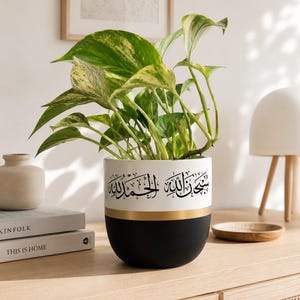 Peut inclure: Un pot de fleurs cylindrique en céramique bicolore, avec une partie supérieure blanche et une partie inférieure noir mat, séparées par une fine bande dorée. La section blanche présente une calligraphie arabe noire. Une plante verte panachée.