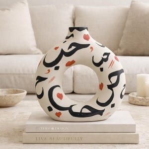 Puede incluir: Jarrón de cerámica crema en forma de donut, adornado con caligrafía árabe negra y motivos de corazones y labios rojos. El jarrón descansa sobre dos libros titulados "INTERIOR DESIGN" y "LIVE BEAUTIFULLY".