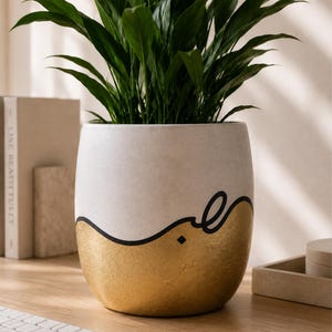 May include: A cylindrical ceramic plant pot featuring a matte off-white upper section and a textured metallic gold lower section, separated by a bold, black, hand-drawn wavy line that forms a stylised Arabic script character in the centre.