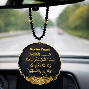 May include: A circular black resin car hanging ornament with gold-colored edges and gold foil accents at the bottom. It features the text 'Dua For Travel' in English and Arabic calligraphy in gold. The ornament hangs from a black beaded chain.