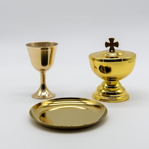 Peut inclure: Ensemble de trois objets religieux dorés : un calice, un bol à couvercle avec une croix sur le dessus et une petite assiette ronde. Les objets sont en métal brillant et sont posés sur un fond blanc uni.