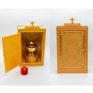 Op de afbeelding: Een goudgeel religieus tabernakel met een kruis erop, open om een gouden kelk te onthullen. De deur aan de rechterkant heeft een kruis en de letters IHS boven een kelk. Een kleine rode kaars staat ervoor.