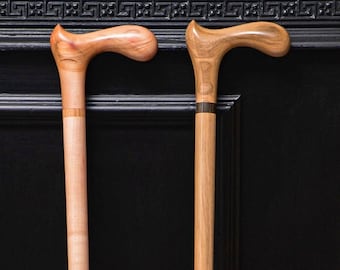 May include: Two wooden walking canes with curved handles. One cane is light-colored with a reddish hue, the other is a lighter brown with a dark band near the top. Both have a smooth finish and are standing upright.