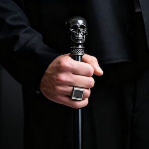 Può includere: Bastone nero con manico a forma di teschio. La persona indossa un abito nero e un anello d'argento con una pietra quadrata nera. Il bastone è tenuto in mano.