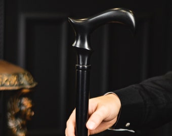 May include: A black wooden cane with a curved handle is held by a person. The cane has a sleek, polished appearance. The background includes a desk lamp with a green shade and a dark bookshelf.
