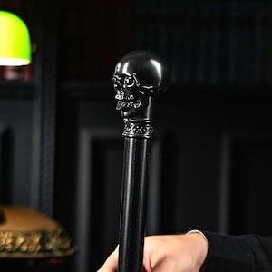 May include: Black skull-topped walking cane. The skull is detailed with eye sockets, a nose cavity, and teeth. The cane's shaft is black and smooth. The cane is held by a person.