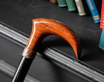 May include: A wooden walking stick with a curved handle. The handle is a rich brown color and has a polished finish. The stick is black and has a silver band near the handle.