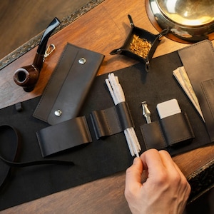 Może przedstawiać: Zestaw skórzanych akcesoriów do palenia fajki, w tym fajka, tacka na tytoń, zestaw do czyszczenia i skórzane etui. Przedmioty są ułożone na drewnianej powierzchni.