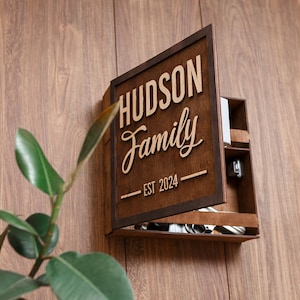 以下が含まれることがあります： レーザーカットされた「HUDSON Family EST 2024」の文字が施された、ダークブラウンの木製壁掛けキャビネット。扉は半開きになっており、内部の収納スペースと金属製の金具、収納されているアイテムが見える状態です。