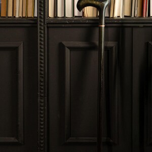 May include: A dark wooden cane with a curved handle and a gold accent ring. The cane stands upright against a black paneled wall with a bookshelf filled with books in the background. The cane is approximately 36 inches tall.
