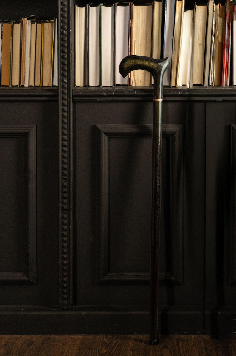 May include: A dark wooden cane with a curved handle and a gold accent ring. The cane stands upright against a black paneled wall with a bookshelf filled with books in the background. The cane is approximately 36 inches tall.