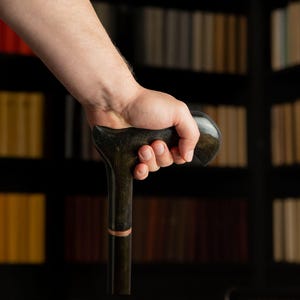May include: A dark wooden cane with a curved handle is held in a hand. The cane has a dark finish with a lighter-colored band near the top. The background features a bookshelf filled with books.