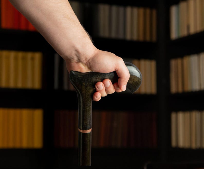 May include: A dark wooden cane with a curved handle is held in a hand. The cane has a dark finish with a lighter-colored band near the top. The background features a bookshelf filled with books.