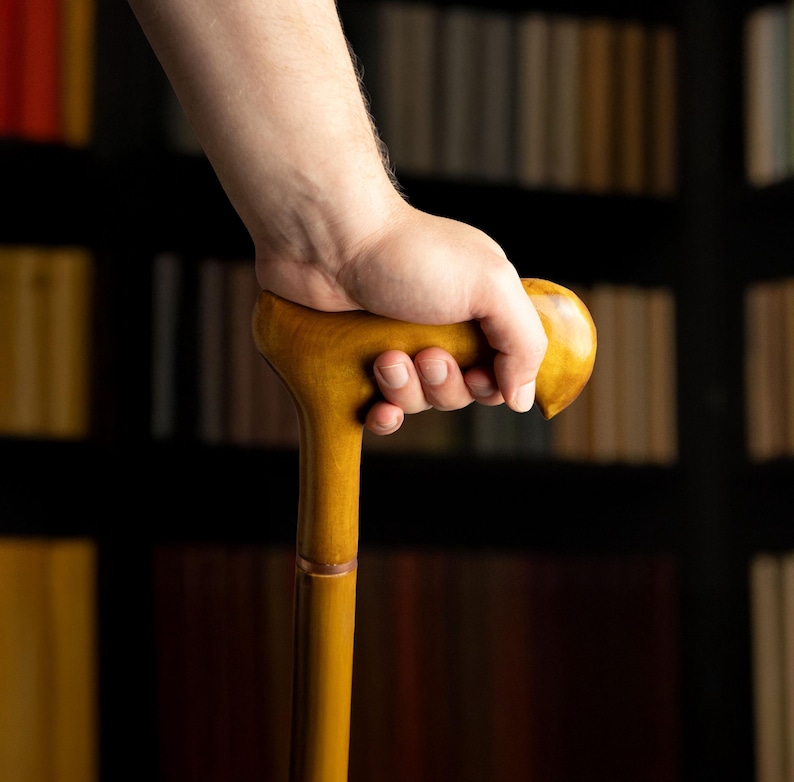 May include: A wooden cane with a rounded handle is held in a hand. The cane is a light brown color and appears to be made of polished wood. The background is a blurred image of a bookshelf.
