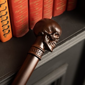 May include: A brown skull-shaped walking stick with a decorative band around the handle. The stick is resting on a dark surface.