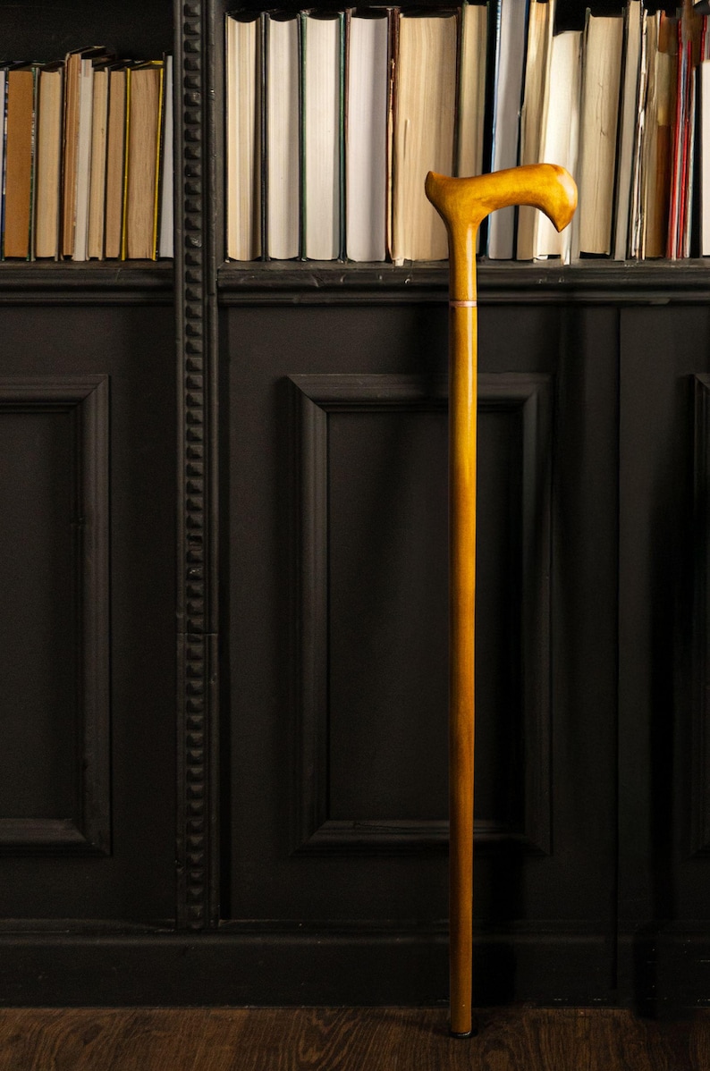 May include: A wooden walking cane with a curved handle, standing upright against a dark paneled wall and a bookshelf filled with books. The cane is a warm, golden color, and the handle is shaped for comfortable gripping. The background features a collection of books.