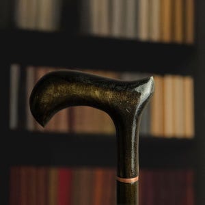 May include: A close-up of a dark wooden cane with a curved handle. The handle has a glossy finish with gold accents. The cane's shaft is dark with a copper-colored ring near the bottom. The background is a blurred bookshelf.