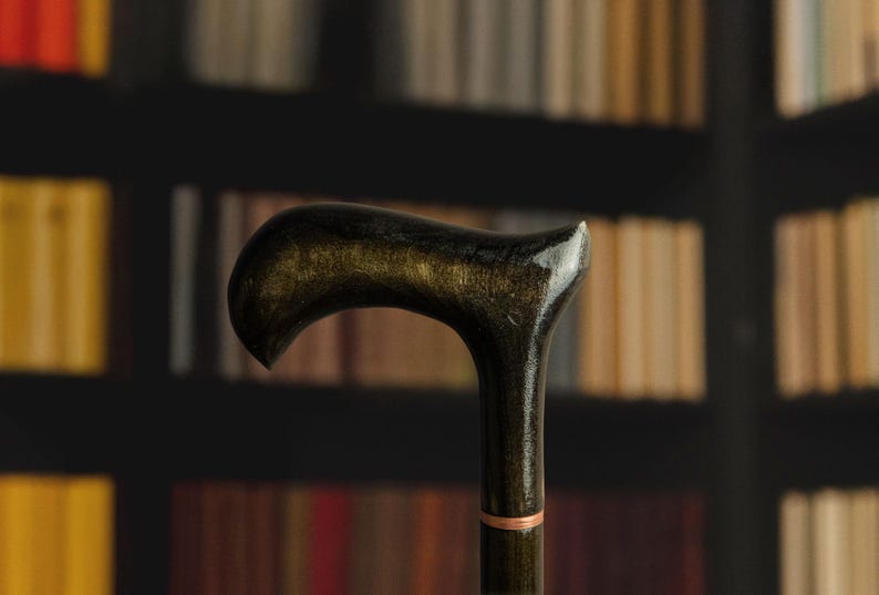 May include: A close-up of a dark wooden cane with a curved handle. The handle has a glossy finish with gold accents. The cane's shaft is dark with a copper-colored ring near the bottom. The background is a blurred bookshelf.
