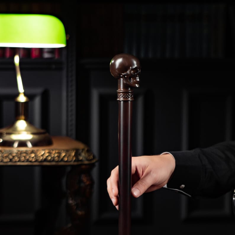 May include: A dark wooden walking stick with a skull-shaped handle. The stick is resting on a table with a green lamp in the background.