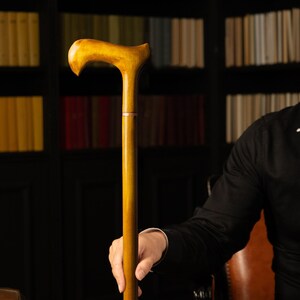 May include: A wooden walking cane with a curved handle and a light brown finish. The cane is held by a person wearing a black shirt. The background features a bookshelf filled with books.