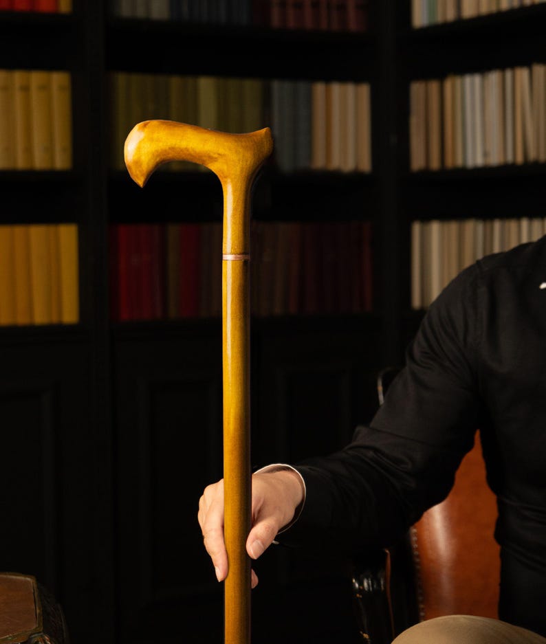 May include: A wooden walking cane with a curved handle and a light brown finish. The cane is held by a person wearing a black shirt. The background features a bookshelf filled with books.