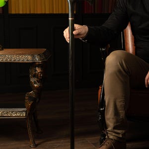 May include: A black wooden cane with a curved handle, accented with gold and a thin pink band. The cane is held by a person in a dark setting, suggesting a classic or vintage style. The cane is approximately 36 inches tall.