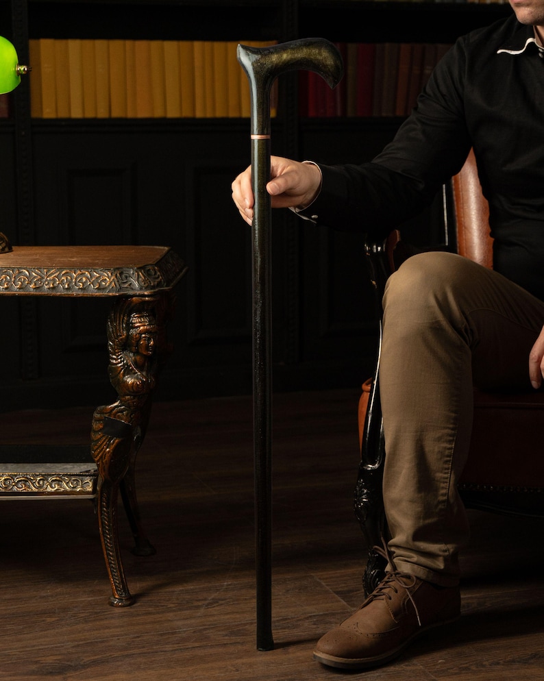 May include: A black wooden cane with a curved handle, accented with gold and a thin pink band. The cane is held by a person in a dark setting, suggesting a classic or vintage style. The cane is approximately 36 inches tall.