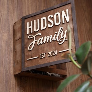 Pode incluir: Uma placa de parede quadrada de madeira com o texto HUDSON Family e EST 2024 em letras claras em relevo sobre um fundo de madeira castanho escuro. A placa é emoldurada por uma borda escura e inclui uma pequena prateleira na borda inferior.