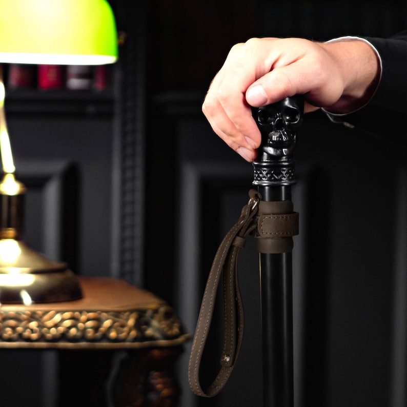 May include: A black walking stick with a skull-shaped handle and a brown leather strap. The handle has intricate metal details.