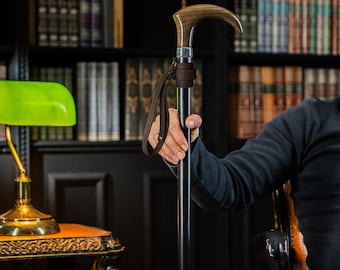 May include: A walking cane featuring a smooth, curved wooden handle with a natural grain pattern, a silver-toned metal collar, and a solid black shaft. A dark brown leather wrist strap is attached to the upper section of the cane just below the handle.