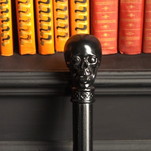 Puede incluir: Un objeto decorativo negro con forma de calavera sobre un poste negro. La calavera tiene rasgos faciales detallados. El fondo incluye una estantería con libros naranjas y rojos, lo que sugiere un tema de Halloween o gótico.
