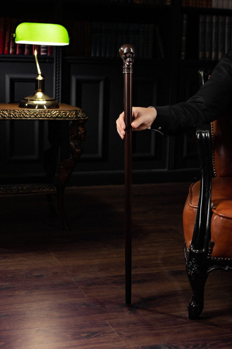 May include: A dark wooden walking stick with a skull-shaped handle. The stick is held by a hand in a dark suit jacket.