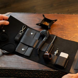 May include: A black leather pipe roll with a tobacco pipe, lighter, and cleaning tools. The roll is open, displaying the items in individual compartments. A small black ashtray sits nearby on a wooden surface.