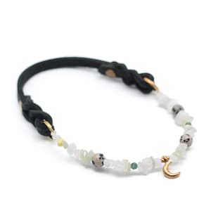 K&ouml;nnte beinhalten: Ein schwarzes geflochtenes Lederarmband mit einem goldenen Halbmond-Anh&auml;nger und wei&szlig;en, gr&uuml;nen und braunen Edelsteinperlen.