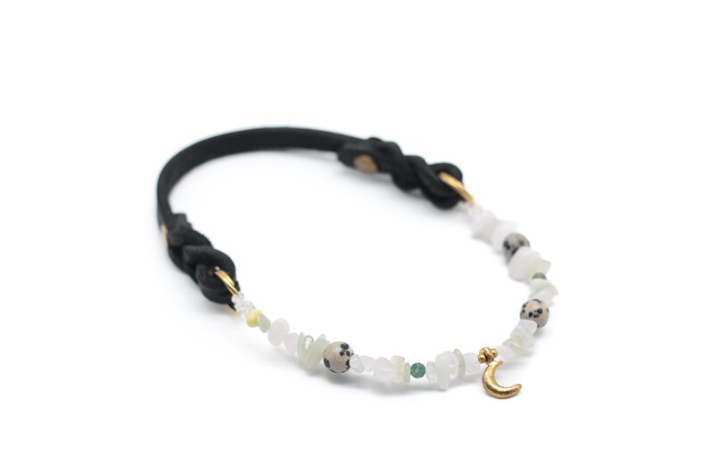 K&ouml;nnte beinhalten: Ein schwarzes geflochtenes Lederarmband mit einem goldenen Halbmond-Anh&auml;nger und wei&szlig;en, gr&uuml;nen und braunen Edelsteinperlen.