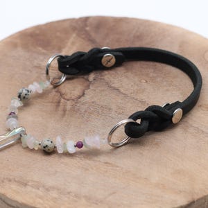 K&ouml;nnte beinhalten: Ein schwarzes Lederhundekragen mit geflochtenem Design und einem silbernen Verschluss. Das Halsband ist mit einem Kristallanh&auml;nger und einer Reihe von wei&szlig;en und rosafarbenen Edelsteinperlen verziert.