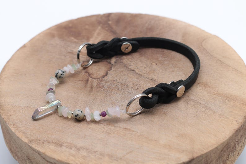 K&ouml;nnte beinhalten: Ein schwarzes Lederhundekragen mit geflochtenem Design und einem silbernen Verschluss. Das Halsband ist mit einem Kristallanh&auml;nger und einer Reihe von wei&szlig;en und rosafarbenen Edelsteinperlen verziert.